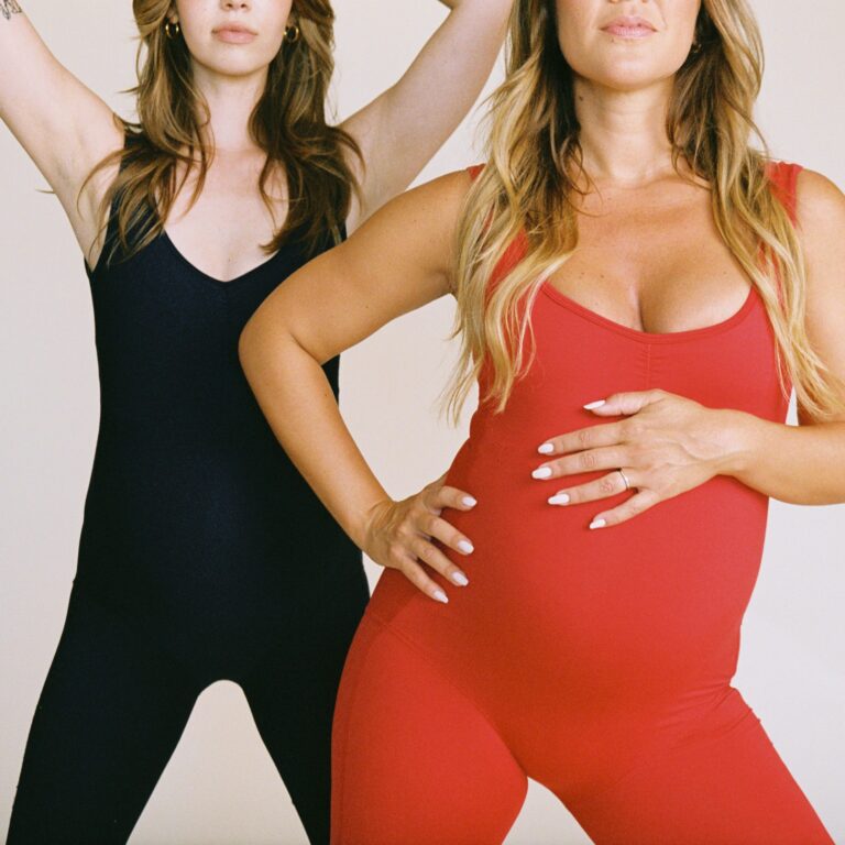 Two women in fitted bodysuits, one black and one red, standing side by side with hands on hips against a neutral background