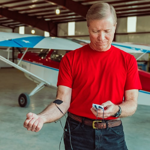 Man in red shirt checking EMF protection device near small aircraft, wrist connected to measurement equipment in hangar