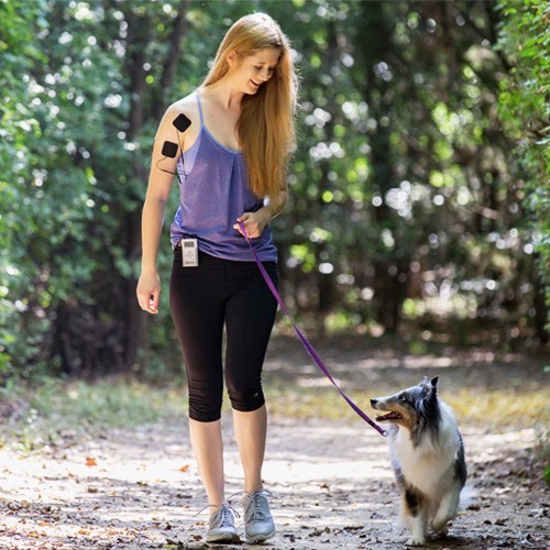 Woman in purple tank top walking dog on forest path, wearing black leggings and carrying SYB EMF protection product on arm