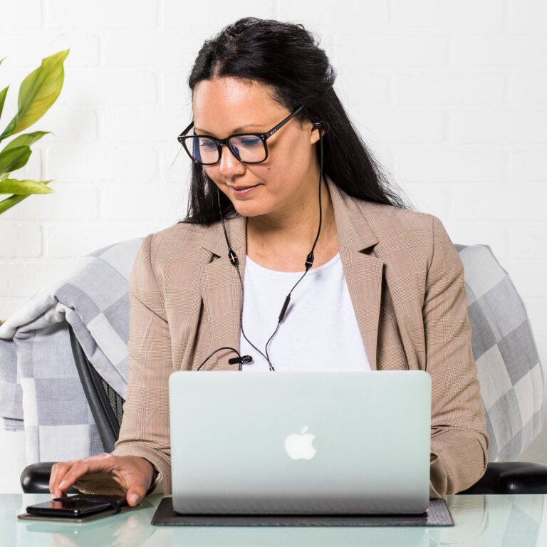 Woman in glasses and beige blazer works on mint green MacBook at desk with plant, focused on screen