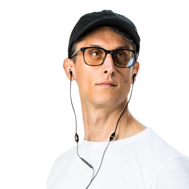 Man in white shirt and black cap wearing glasses and earbuds, looking slightly to the side against white background