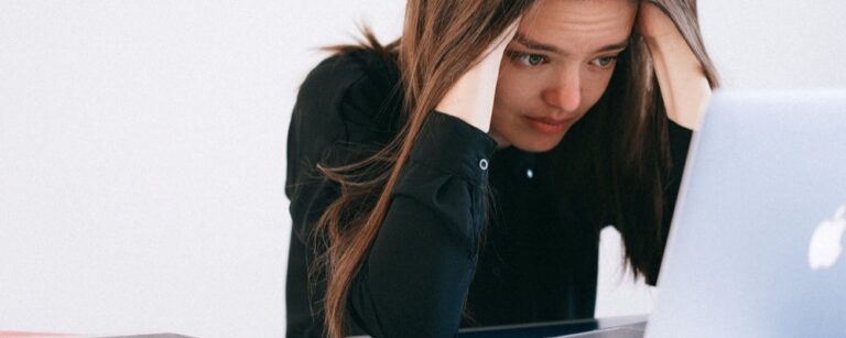 Woman in black sweater looking stressed, hand on head, working at white laptop
