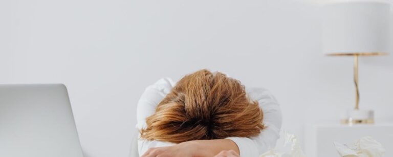Person with brown hair slumped forward at desk with laptop, suggesting fatigue or stress