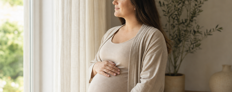 Pregnant woman in a softly-lit modern home, hand on belly, looking at natural light through a window